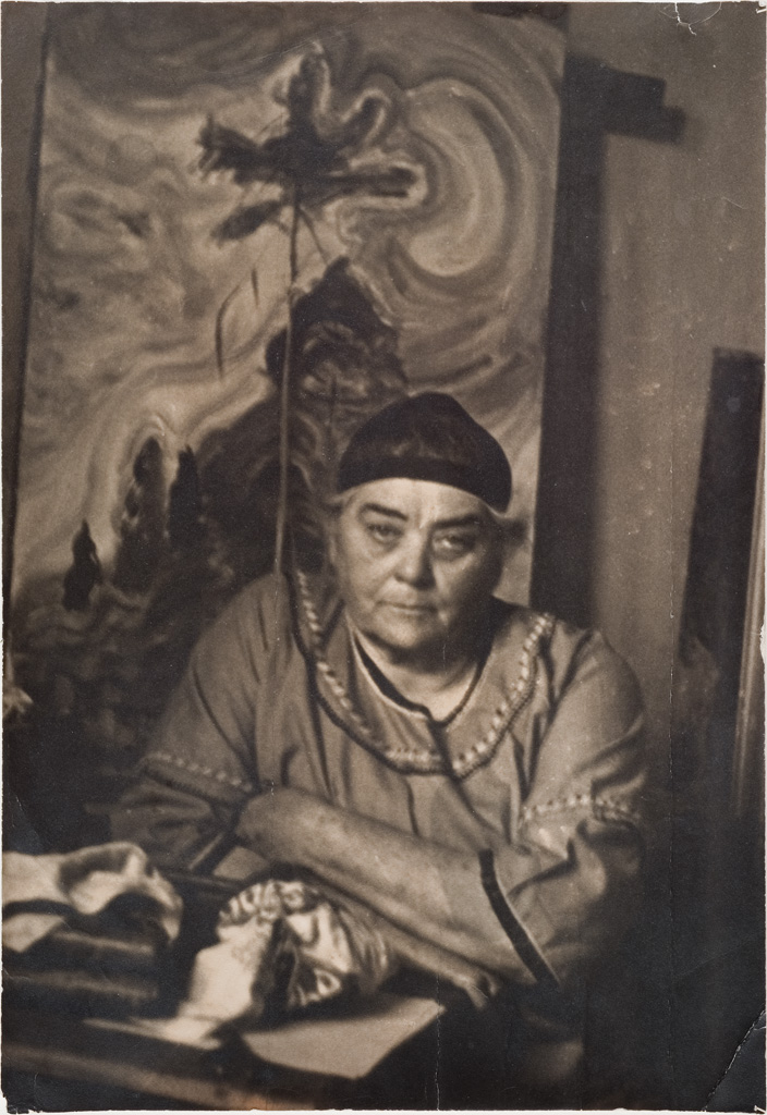 Sepia-toned photograph of Emily Carr in her studio with one of her paintings depicting a big tree in the background.