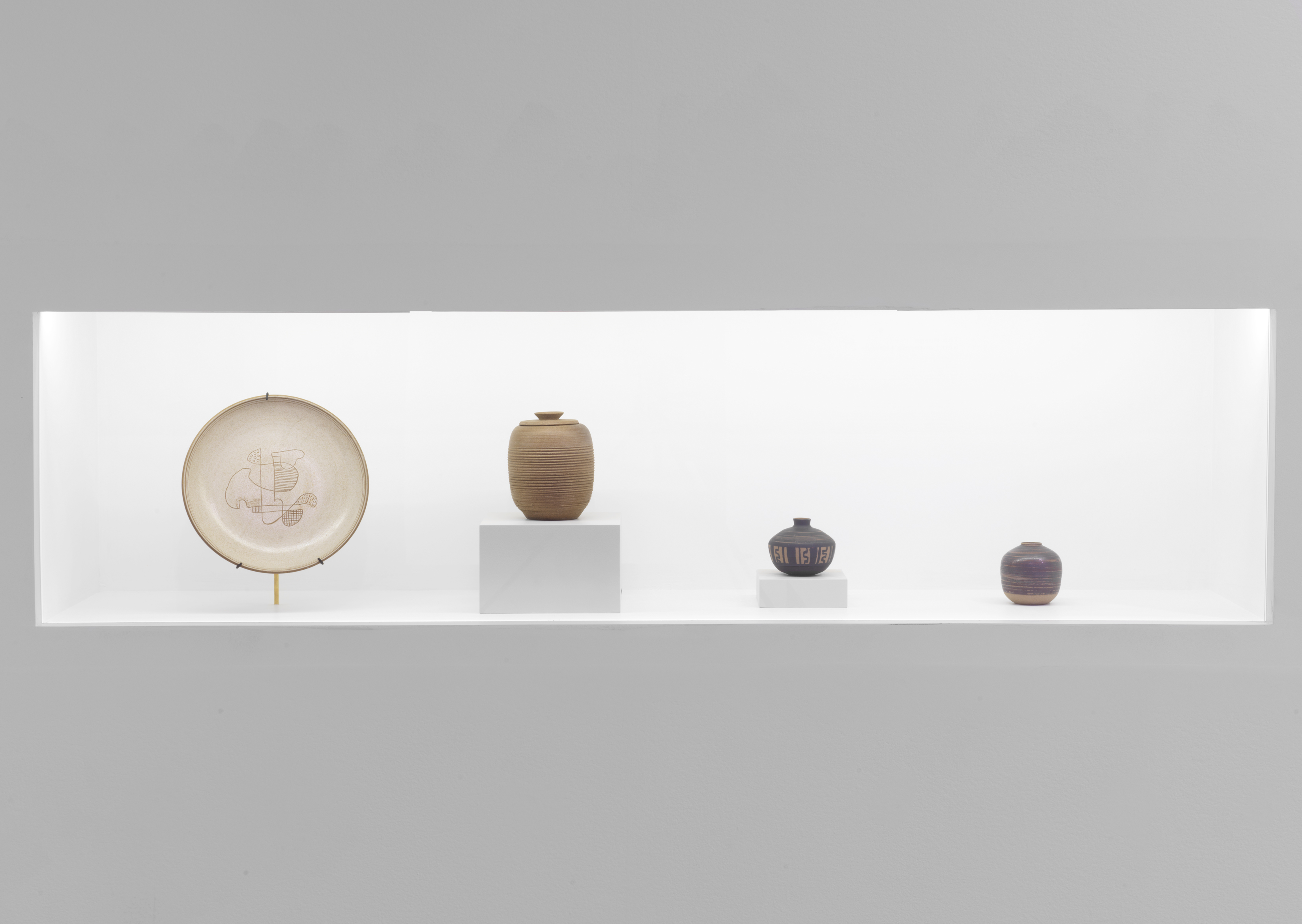 Photograph displaying four ceramic pieces, from left to right: one beige plate a brown vase on top of a white cube, a blue and brown vase and a smaller purple and beige vase, all of different sizes.