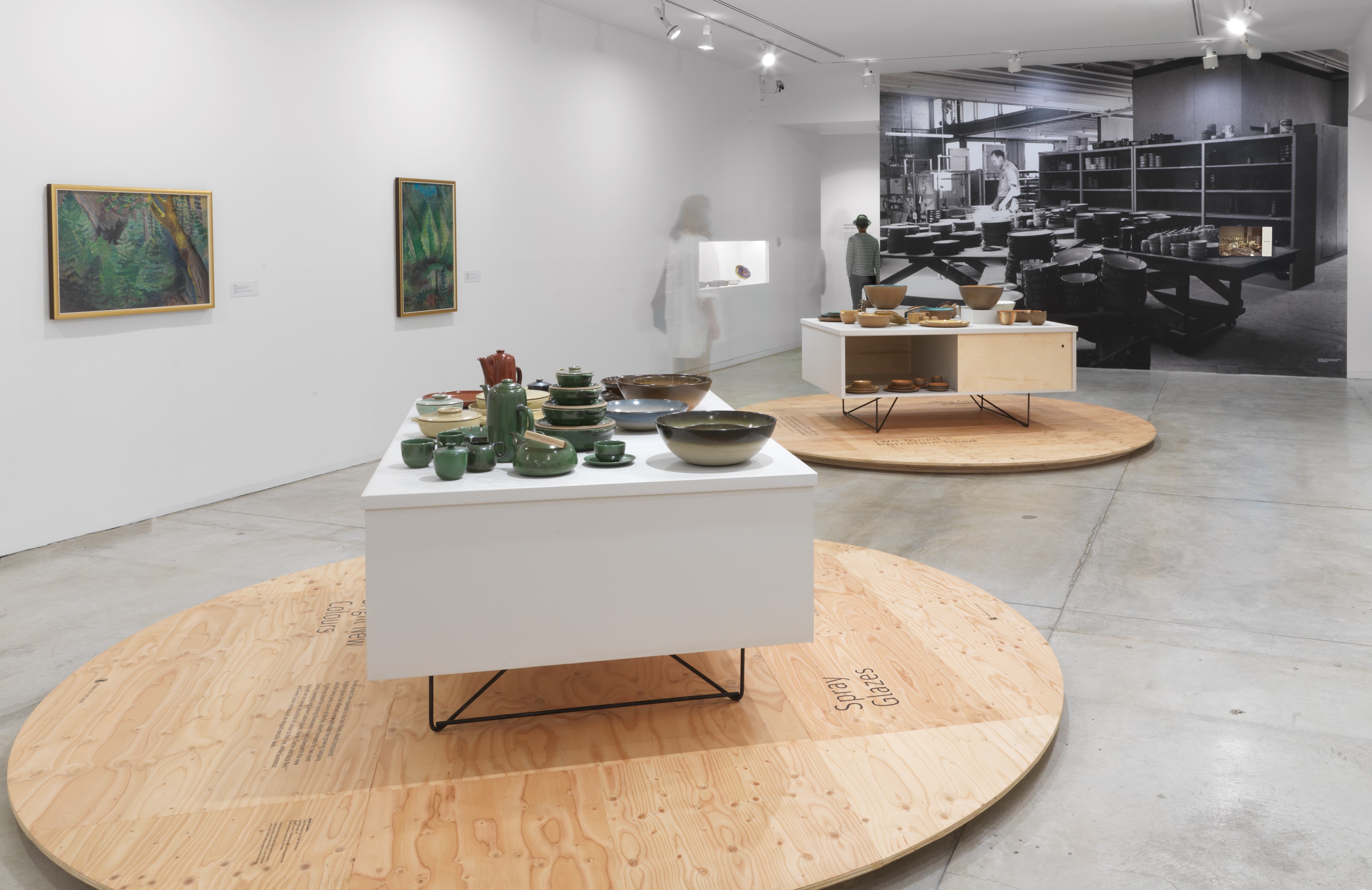 Installation view of the exhibition, with a black and white mural in the background and two cabinets holding ceramic pieces in several colours.