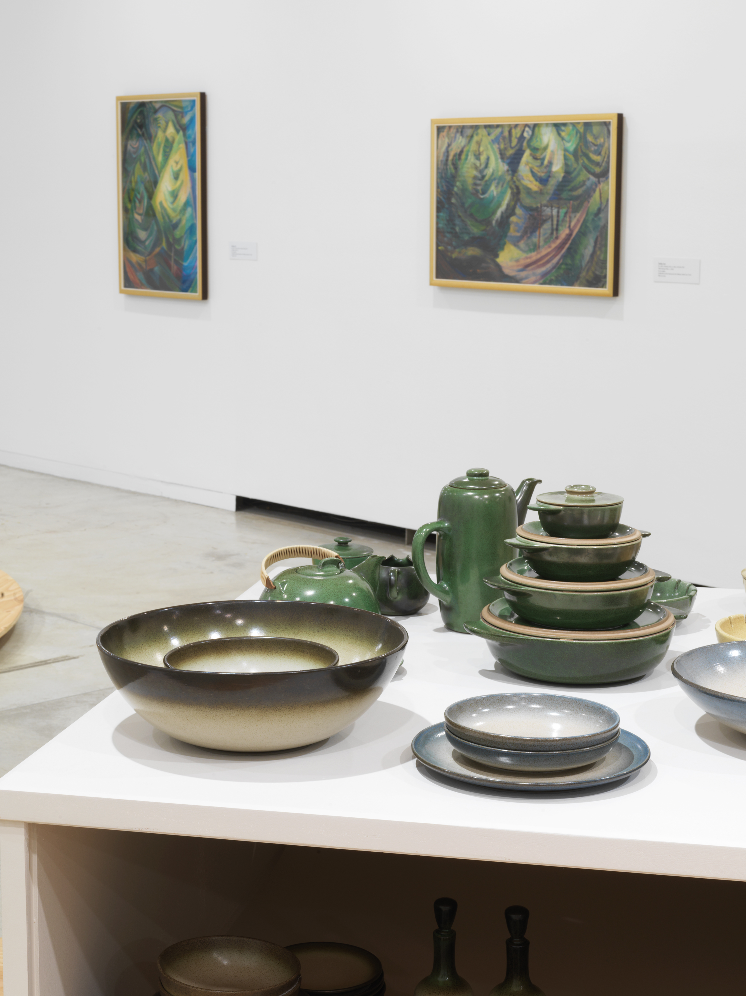 Ceramic bowls, plates and teapots in blue, green and browns hues, with two paitings by Emily Carr, depicting forests scenes in green and brown colours in the background.