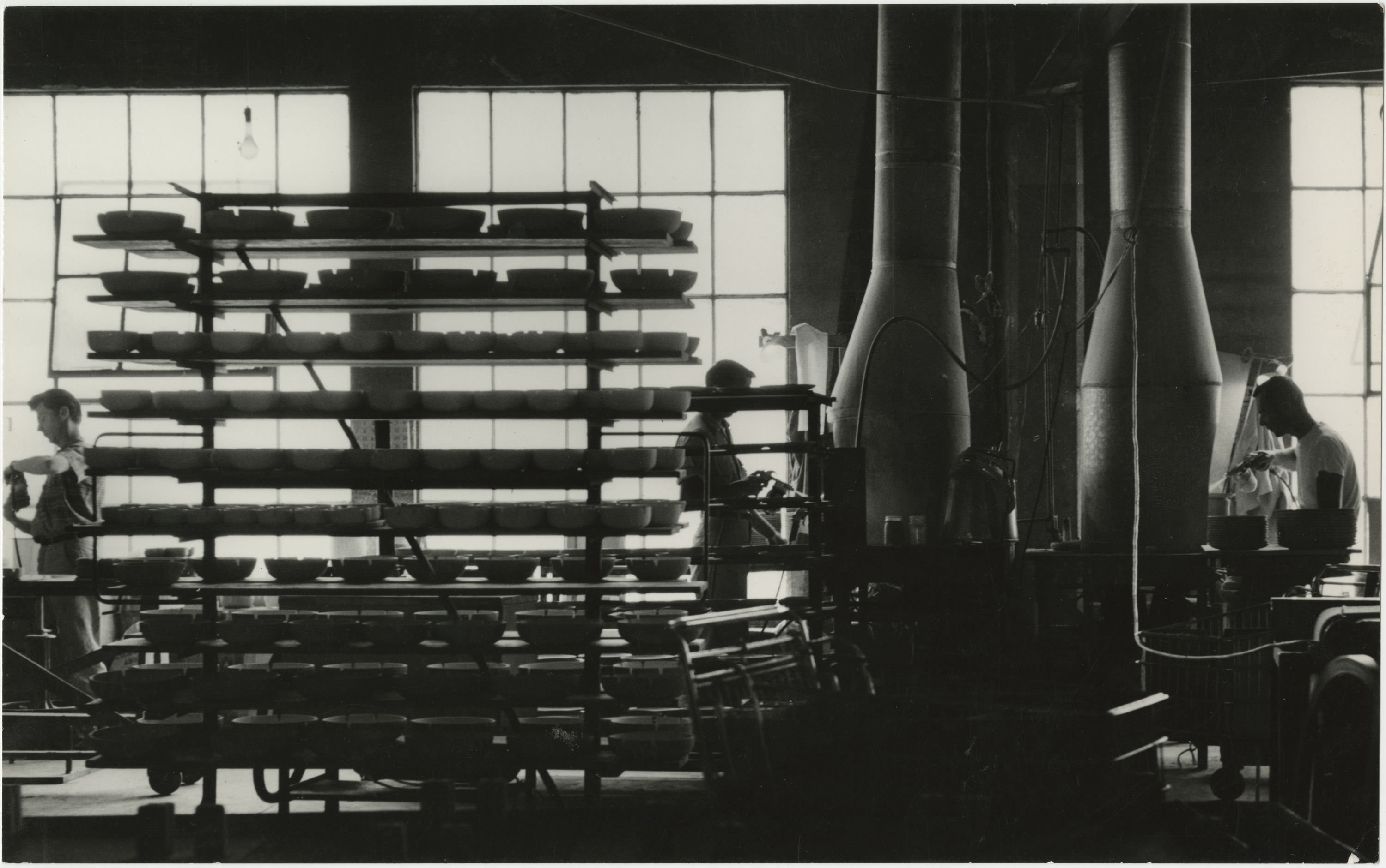 Black and white photo of men working in the background and Heath ashtrays on racks.