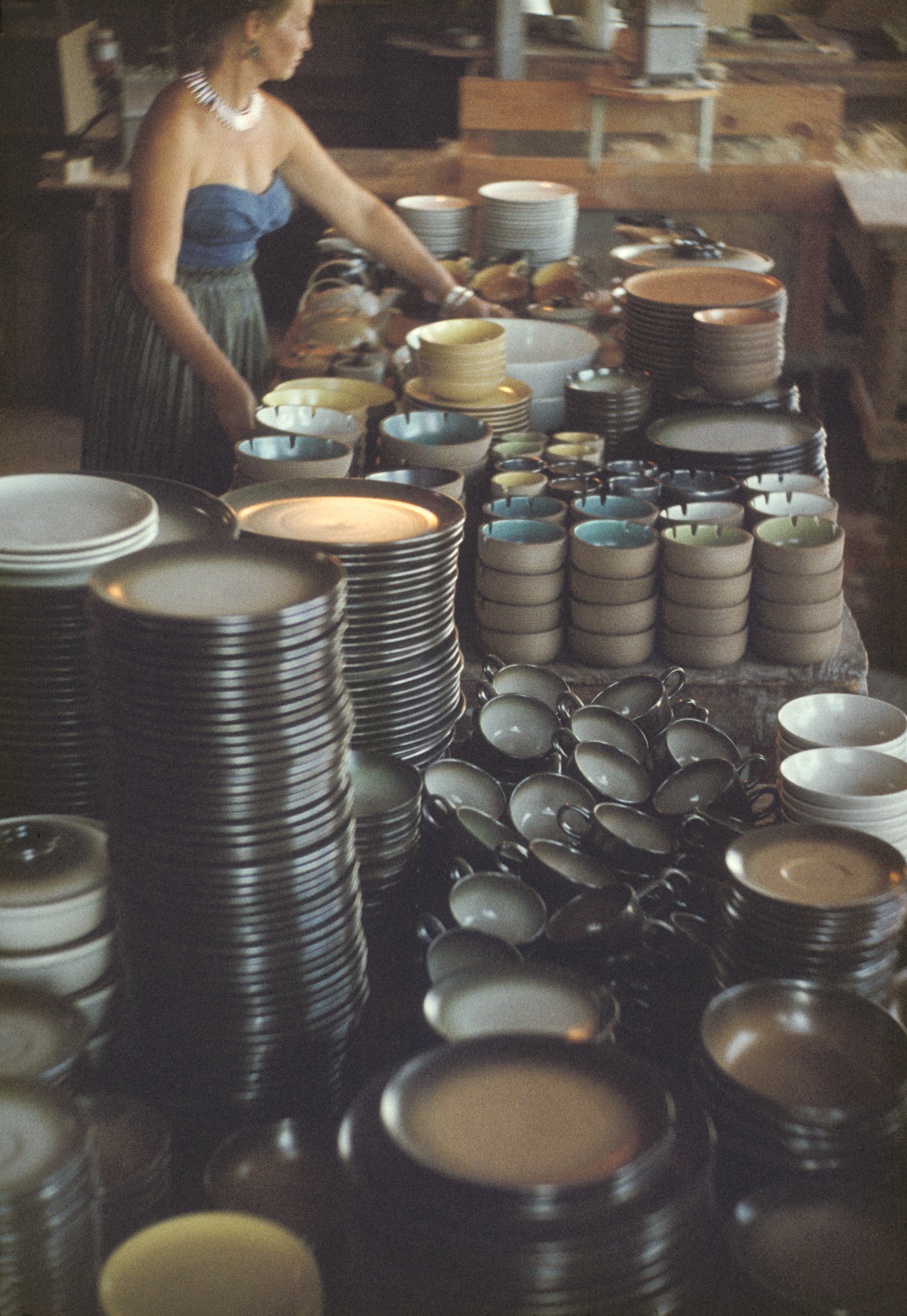 A woman in a dress organizing stacks of plates, cups, ashtrays and bowls on a long table.