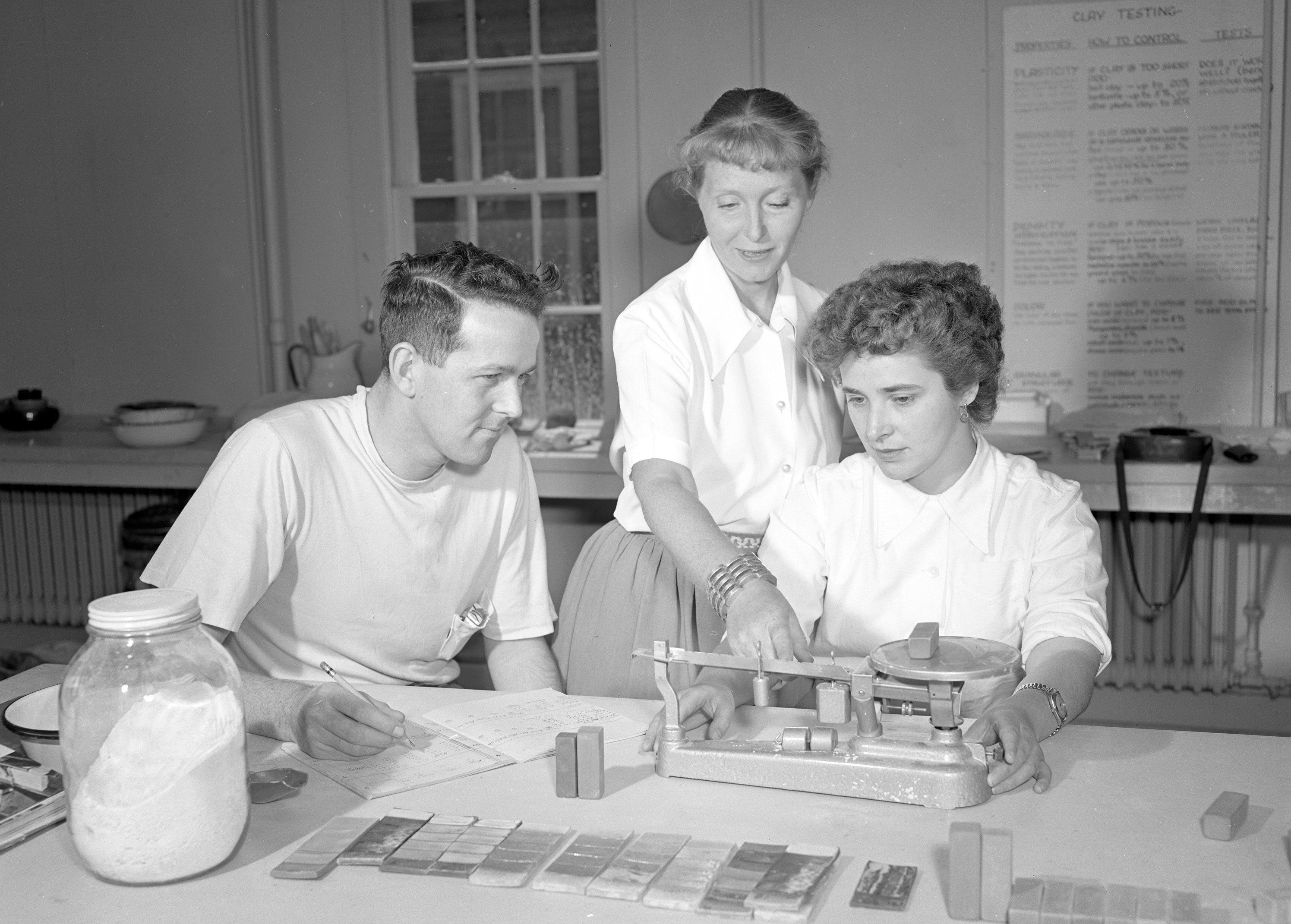 Black and white photograph of three people, one standing in between the other two, in front of a table with ceramic pieces on it.